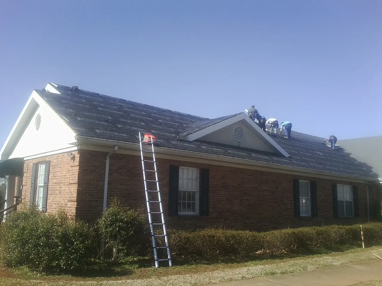 Crew tearing shingles off a brick ranch roof with a ladder set — Emergency Roof Repair work by our crew in Fitzwilliam
